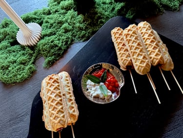 Golden waffle corn dogs on wooden skewers served on a black platter with dipping sauces.