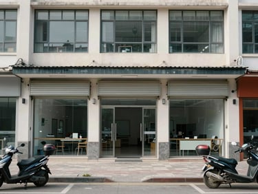 Street-level exterior of a standalone commercial building, ground floor shutters open showing interior depth, upper floors with office windows, overcast midday light, pavement and parked scooters in foreground for scale, no people, documentary framing