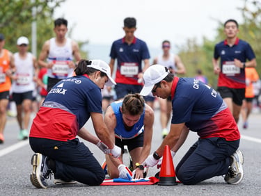 Volunteers in Croix Blanche uniform providing first aid at a marathon, focused on a runner, natural overcast daylight, crisp navy and signal red accents, sharp focus, 35mm lens.