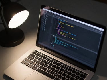 Close overhead shot of a laptop screen showing clean HTML and CSS code in a dark editor, hard tungsten lamp light from upper-left, sharp contrast, no reflections, matte desk surface visible at edges