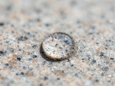 Extreme close-up macro shot of a liquid droplet perfectly beading up on a sealed light-colored granite surface, showing high surface tension, bright daylight, razor-sharp focus.