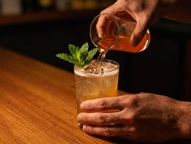 Hands pouring a vibrant cocktail with fresh mint garnish, warm low-key evening light, shallow depth of field, 35mm film.