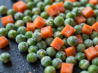 Close-up of vibrant, IQF-frozen green peas and diced carrots scattered on a frosted dark surface, capturing the texture and icy sheen.