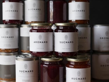 Stack of elegant, minimalist branded food cans and a glass jar of fruit pulp, with soft studio lighting highlighting the labels.