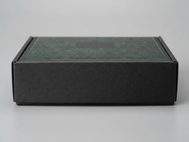 Macro shot of a pristine, empty rigid cosmetic box showing crisp edges, matte charcoal texture, and precise forest green foil stamping, soft diffused studio lighting, neutral minimalist backdrop, 85mm