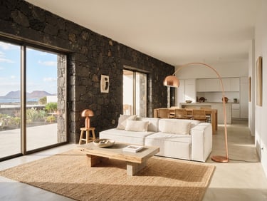 Minimalist open-plan living room in Lanzarote, raw volcanic stone wall contrasting with crisp white plaster, warm natural sunlight, copper floor lamps, editorial architectural photography