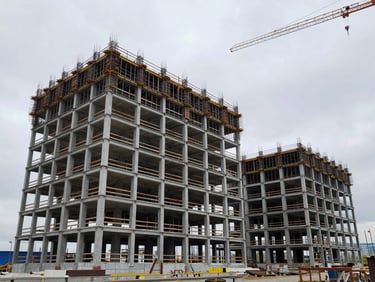 Wide shot of a commercial building under construction in Novosibirsk, steel skeleton and cladding panels being installed, overcast sky, crane visible at upper right, documentary framing