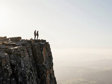 A couple standing on a dramatic cliffside, backlit by harsh light, massive negative space.