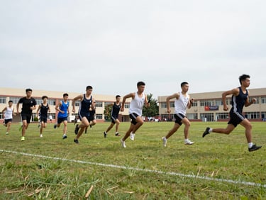 Wide shot of students running relay races on a grass field, bright overcast daylight, school building visible in background, candid motion blur on running feet