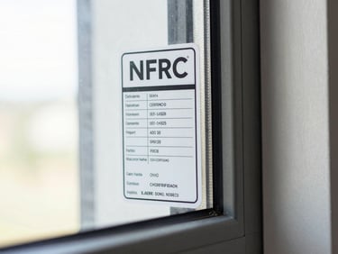 Close-up of an NFRC certification label affixed to a vinyl window frame, sharp focus on printed ratings text, clean white background behind the frame, bright even lighting