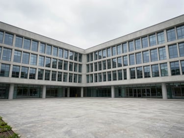 Wide exterior shot of a modern university campus building in flat overcast daylight, clean concrete and glass facade, empty courtyard, no people posing