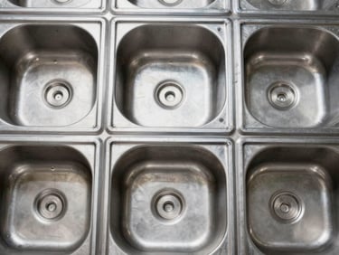 Tight overhead shot of stainless steel sink blanks arranged in a factory grid, studio strobe, even industrial lighting, showing gauge thickness and weld seams, no warmth or atmosphere
