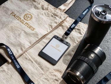 Tight overhead shot of custom branded merchandise — embroidered tote bag, lanyard, and tumbler arranged on a dark surface under north-facing daylight, close crop revealing stitch texture and foil detail on each piece