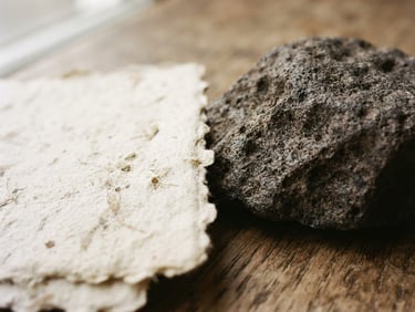 Macro photograph of textured handmade paper and raw dark stone under soft natural window light, warm editorial style, 35mm