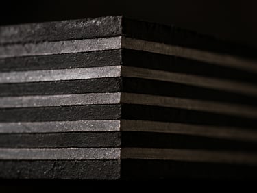 Extreme close-up cross-section of a laminated neoprene elastomeric bearing pad cut cleanly to reveal alternating black rubber layers and bright steel shim plates, harsh industrial workshop lighting from the side casting sharp shadows that emphasize the precision of each laminate layer, raw cut surface showing material density and bonding quality