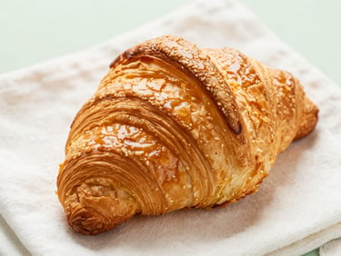 Close-up of a golden, flaky croissant showing immaculate lamination, resting on a clean linen cloth under natural light.