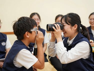 A child laughing during a friendly eye screening with a portable camera held by a smiling volunteer wearing a navy vest, warm indoor light