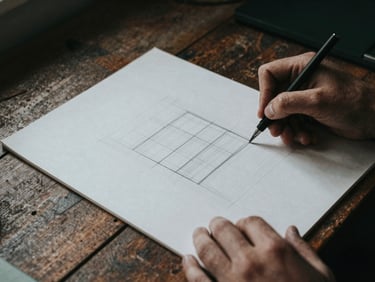 Hands sketching a wireframe layout on a heavy paper pad on an industrial wooden desk, moody natural light, high contrast.