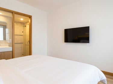 Premium double bed AC room, spotless white sheets, flat-screen television on wall, modern attached bathroom visible