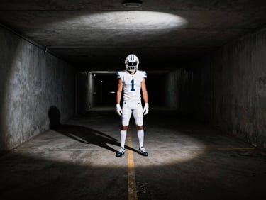 High-contrast wide shot of a football player standing in a dark, empty stadium tunnel under a single harsh white spotlight, heavy shadows.