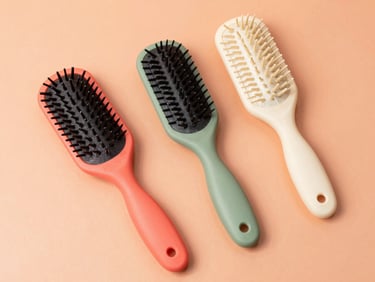 Clean overhead flat-lay of three curl-defining brushes in coral, sage, and cream colors arranged on a bright peach backdrop, sharp focus.