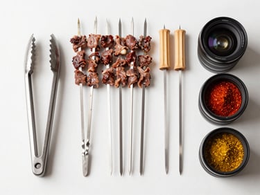 Bright overhead view of a braai accessory display — tongs, skewers, firelighters, and spice rubs arranged neatly on a clean white surface under studio light