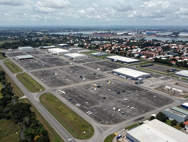 Elevated wide-angle aerial photograph of a 200-hectare industrial logistics platform in Apartadó, Antioquia, Colombia, shot from above looking south-east, paved internal roads forming a grid pattern across the site, warehouse pads and utility corridors clearly delineated, Route 62 highway visible along the left edge, port infrastructure on the far horizon, overcast natural daylight for maximum detail clarity, no people, no vehicles