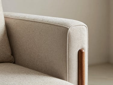 Close-up of a quality upholstered chair arm showing stitched fabric detail and solid wood leg, natural daylight, clean neutral background wall visible