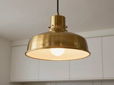 Detail of a designer pendant lamp in brushed brass over a kitchen island, warm tungsten light glowing inside the shade, plaster ceiling and white cabinetry visible
