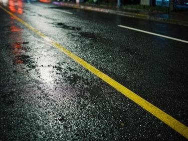 Tight, atmospheric crop of wet asphalt with yellow lane markings reflecting moody night-time streetlights, cinematic film grain, 35mm lens