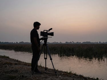 Documentary camera on a tripod at the edge of a haor embankment at dusk, operator's silhouette visible against a fading amber-grey sky, water and reed beds stretching behind, natural available light only