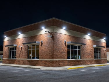 Exterior of a commercial building at night, multiple cameras visible on the facade, cold security lighting casting sharp shadows on the brick, empty parking lot in foreground
