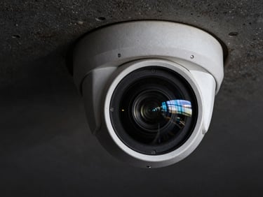 Extreme close-up of a dome surveillance camera lens mounted on a dark concrete ceiling, glass lens reflecting a faint blue-tinted light, deep black background, sharp hardware detail