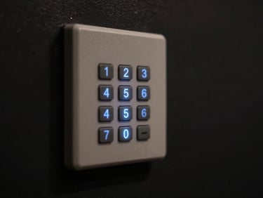 Close macro of a keypad alarm panel glowing faintly in a dark hallway, backlit buttons casting soft blue light on the surrounding wall, near-black background