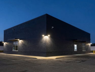 Wide shot of a dark commercial building exterior at night, security cameras visible on corner brackets, blue-hour sky, empty parking lot, low ambient lighting emphasising building scale