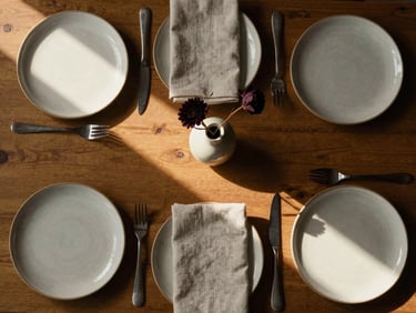 Overhead flat-lay of a private dinner table set for six, artisan ceramic plates, hand-folded linen napkins, a single dark floral stem in a slim vase, warm side-light casting long shadows across the table surface