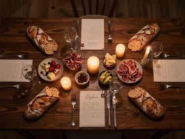 Overhead flat-lay of a private dinner table for eight, dark wood surface, hand-written place cards, single taper candles, artisan bread and charcuterie arranged with deliberate asymmetry, warm amber candlelight glow