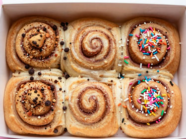 A box of four assorted creative cinnamon rolls with colorful toppings like crushed cookies and sprinkles, flat lay