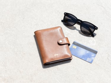 A flat-lay of travel essentials including a leather passport holder, sunglasses, and a credit card on a clean stone surface, bright daylight, 35mm