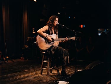 Cinematic wide shot of a folklore artist playing an acoustic guitar on a dark stage, high-contrast warm copper spotlighting, deep shadows, 35mm photography