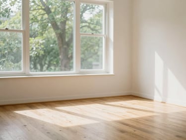 Empty sun-lit room with clean hardwood floors, large windows showing soft green trees outside, pristine white walls, warm editorial feel