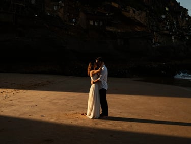 Intimate portrait of a couple, Mariana and João, embracing on a dramatic Portuguese beach at golden hour, low sun creating rim light, high contrast, cinematic wedding photography, deep shadows, warm golden accents, rich contrast, 35mm film grain, wide shot.