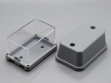 Side-by-side technical isometric shot of two thermoplastic enclosures on a gray surface, one with a clear transparent cover showing internal DIN rail, one with a solid opaque gray cover, high clarity.