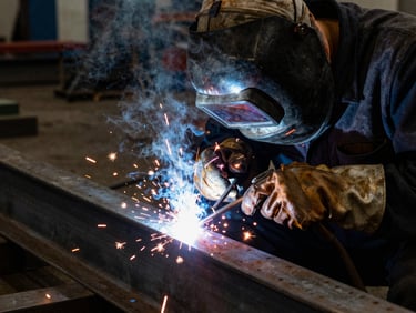 Close-up of a welder in full PPE, actively arc welding a thick steel beam, intense sparks and smoke, high-contrast industrial lighting, deep shadows, cool steel tones with warm amber highlights, 35mm.