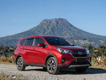 Environmental shot of a red Perodua Ativa SUV parked on a gravel roadside near the foothills of Mount Kinabalu, clear blue sky, bright natural daylight.