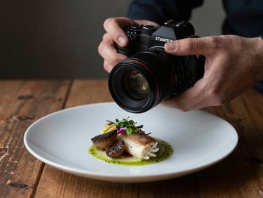 Close-up shot of hands adjusting a camera lens over a freshly plated artisanal dish on a rustic wooden table, soft Scandinavian side-light, moody shadows.