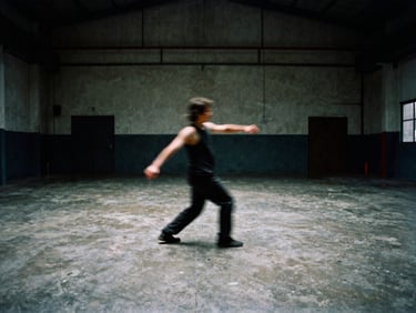 A performer moving dynamically in an industrial warehouse space, long exposure motion blur, raw concrete floor, high-contrast ambient light, 35mm film grain