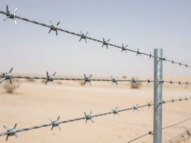 Close-up of high-quality galvanized steel barbed wire tensioned over a secure fence post, sharp metallic details, desert background, bright daylight, professional industrial photography, 35mm