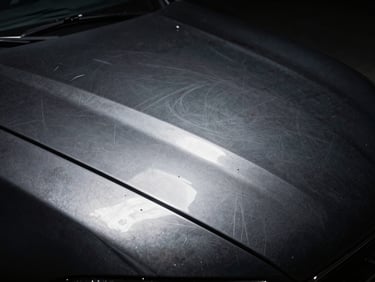 Macro shot of a black car hood under a single point light source showing heavy swirl marks, spiderweb scratches, and dull oxidized paint, harsh contrast