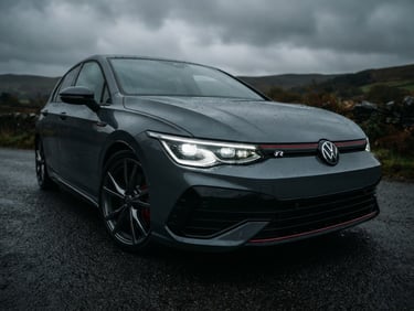 Low-angle close-up of a clean dark metallic grey German hatchback under moody overcast South Wales skies, sharp LED headlights illuminated, clean background